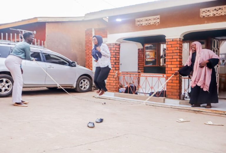 Three women play skipping rope outside a brick house; one jumps while the others turn the rope and smile.