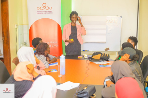 A facilitator stands and talks to a group of women seated around a table, next to the RMHN banner.